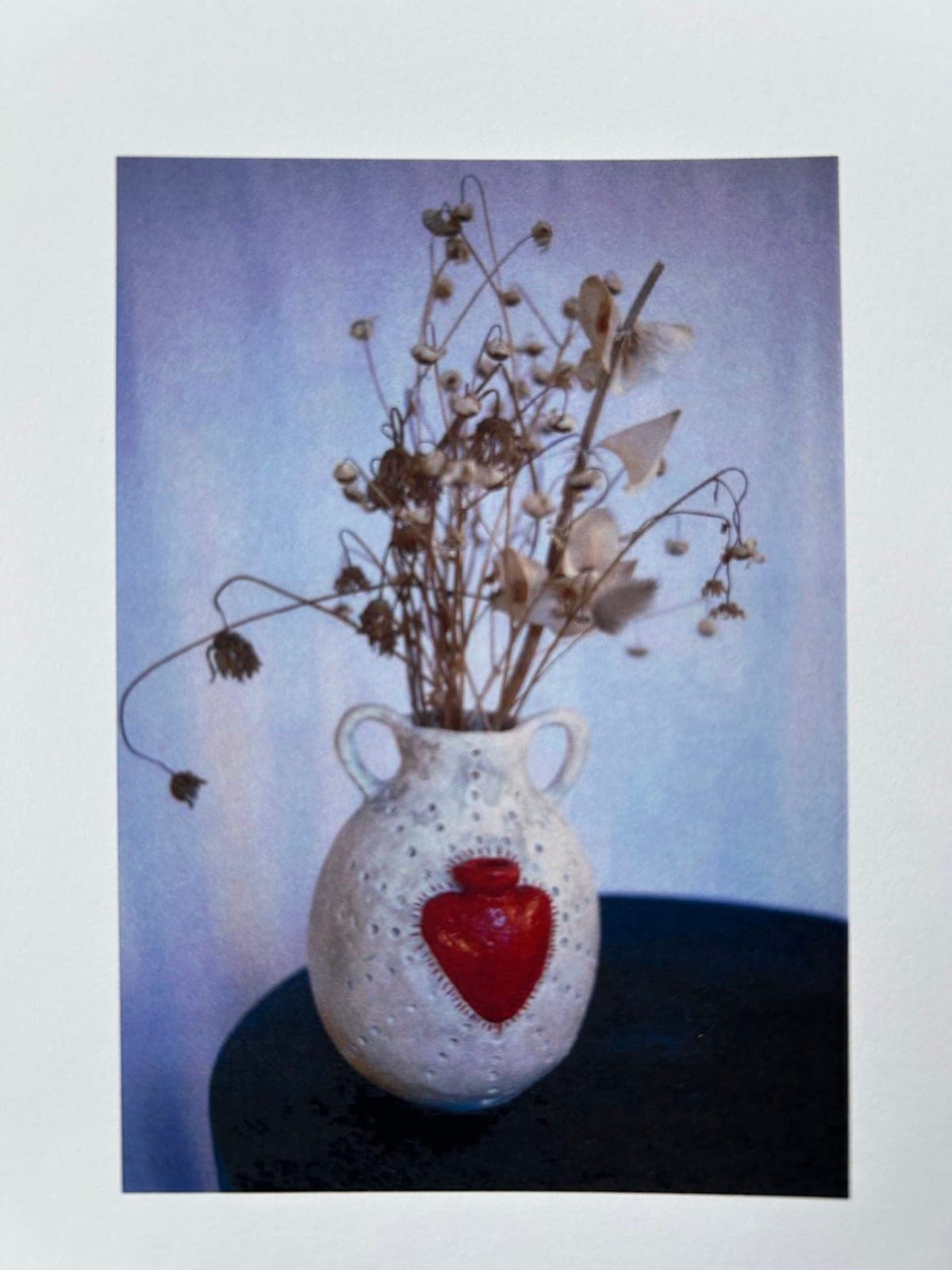 A white speckled vase with a raised red heart, holding dried flowers
